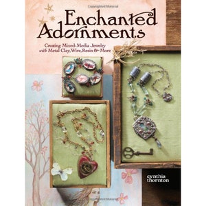 Op de afbeelding: Een boek getiteld "Betoverende Sieraden: Het maken van mixed-media sieraden met metaalklei, draad, hars en meer" door Cynthia Thornton. De omslag toont drie sieraden die worden gepresenteerd in houten dozen met een groene stoffen voering. De sieraden omvatten een ketting met een hartvormige hanger, een ketting met een hanger in de vorm van een medaillon en een armband met verschillende bedels.