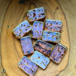 May include: A collection of ten rectangular glass beads with a translucent, opalescent finish. Each bead features an irregular, recessed pattern filled with a dark, metallic copper-toned substance, creating a contrast against the pale, milky surface.