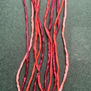 May include: A collection of thin, flexible fabric cords in varying shades of red, including deep burgundy, bright crimson, and soft coral pink. The cords have a smooth, slightly lustrous texture and are arranged in vertical, overlapping strands.