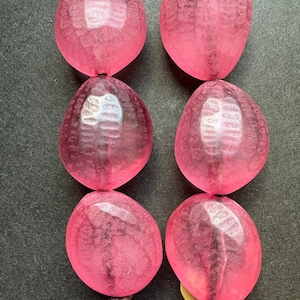 May include: Two vertical strands of translucent, hot pink, pebble-shaped beads. Each bead features a textured, dimpled surface pattern. The beads are strung on dark brown cord, with a small yellow accent visible at the base of the right strand.