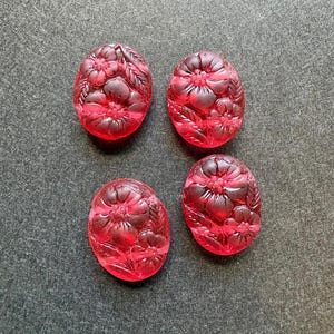 Puede incluir: Cuatro cabujones de vidrio rojo translúcido de forma ovalada con un relieve detallado de flores y hojas. Cada pieza muestra una superficie lisa y redondeada con intrincados patrones florales grabados en el material rojo vibrante.