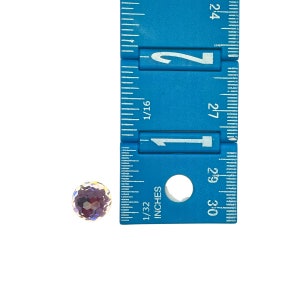 K&ouml;nnte beinhalten: Nahaufnahme eines kleinen, runden, regenbogenfarbenen violetten Edelsteins mit einem Durchmesser von etwa 1,25 cm. Der Edelstein liegt auf einer wei&szlig;en Oberfl&auml;che neben einem blauen Lineal.