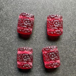May include: Four rectangular, translucent red beads featuring a carved floral pattern on the surface. Each bead displays a detailed relief of flowers and leaves, with a smooth, rounded edge design. The beads are arranged in a two-by-two grid formation.