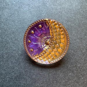 May include: A circular button featuring a split design with one side in deep purple displaying a floral-like pattern with small gold dots and the other side in amber yellow with a honeycomb texture, all enclosed by a metallic twisted rope border.