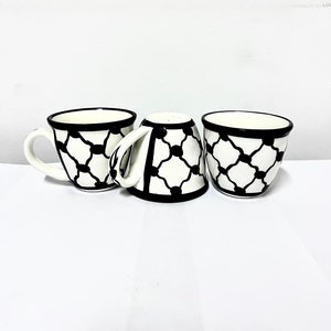 Könnte beinhalten: Drei weiße Keramik-Kaffeetassen mit schwarzen geometrischen Mustern. Die Tassen haben eine abgerundete Form und einen kleinen Henkel.
