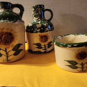 May include: Set of three ceramic containers with a sunflower design. The containers are glazed in a green and white speckled pattern. The largest container is a pitcher with a handle. The middle container is a smaller pitcher with a handle. The smallest container is a bowl.