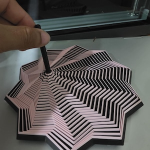 May include: A multi-layered, star-shaped geometric object featuring alternating black and light pink concentric rings. A thin, black cylindrical rod is positioned vertically in the centre of the structure, extending upwards from the core of the design.