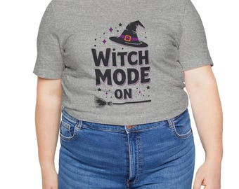 Può includere: Una maglietta grigia melange con un cappello da strega nero e il testo "Witch Mode On" in nero. Il cappello ha una fascia viola e una fibbia nera. Ci sono piccole stelle viola intorno al cappello.