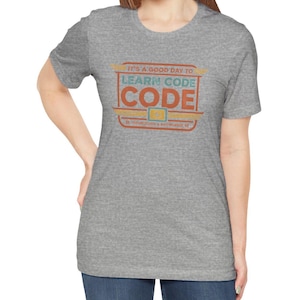 May include: A heather gray short-sleeve t-shirt featuring a rectangular graphic with the text IT'S A GOOD DAY TO LEARN CODE, the word CODE in large orange letters, and the phrase TECHNOLOGY & KNOWLEDGE at the bottom in a retro-inspired design.
