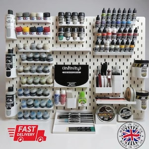 May include: A white pegboard with a collection of model painting supplies, including paint bottles, brushes, and masking putty. The image features the text "(infinity)" and "MADE IN THE UK". The display includes a variety of colours and tools for miniature painting.