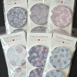 May include: A collection of car coasters with various floral and leaf patterns in shades of purple, blue, and pink. Each coaster is packaged in a clear plastic sleeve with a label that reads "Car Coasters, Handmade with love."
