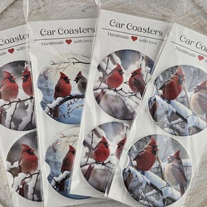 May include: Several car coasters are displayed, each packaged in clear plastic. The coasters feature a winter scene with red cardinals perched on snow-covered branches. The packaging reads "Car Coasters" and "Handmade with love."