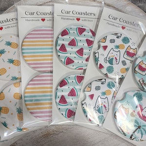 May include: Set of eight car coasters with various summer-themed designs. The designs include pineapples, watermelon, palm trees, and cats on a flamingo float. The coasters are white with colorful designs and the text "Car Coasters Handmade with love" is printed on each card.