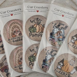 May include: A collection of car coasters with fall-themed designs. Each coaster features a unique illustration, including pumpkins, trucks, and gnomes. The coasters are packaged in clear plastic and displayed in a wooden tray.
