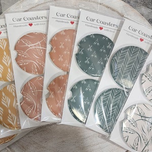 May include: Set of six car coasters with various patterns. The coasters are round and feature a white background with different colored designs. The designs include a chevron pattern, a leaf pattern, and a geometric pattern. The coasters are packaged in clear plastic bags with a white label that reads "Car Coasters Handmade with Love".