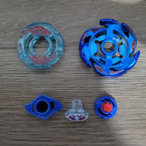 May include: A disassembled blue and clear toy, likely a spinning top, with various components. The parts include a blue top with red accents, a clear ring with red details, and smaller blue and clear pieces. The items are arranged on a wooden surface.