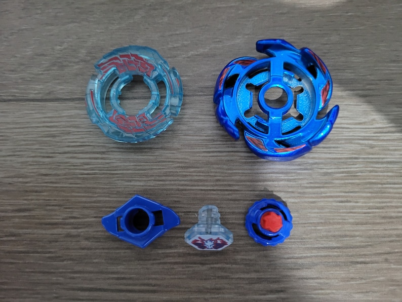 May include: A disassembled blue and clear toy, likely a spinning top, with various components. The parts include a blue top with red accents, a clear ring with red details, and smaller blue and clear pieces. The items are arranged on a wooden surface.
