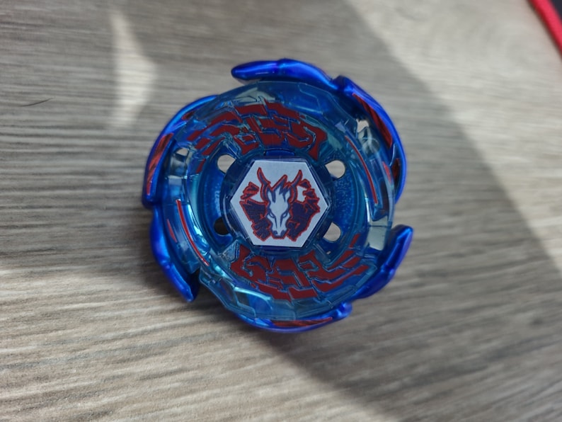 May include: A blue and red spinning top toy with a central emblem. The toy has a circular design with red accents and a central emblem featuring a stylized animal. The toy is designed for spinning and battling.
