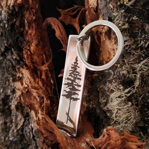 May include: Silver keychain with a tree design engraved on a rectangular tag. The keychain has a silver ring.