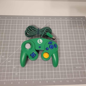 Puede incluir: Mando de videojuegos verde con cable verde. El mando tiene un joystick gris, botones azules y amarillos y una cruceta direccional verde. La letra "L" está en el mando. El cable mide aproximadamente 152 cm de largo.