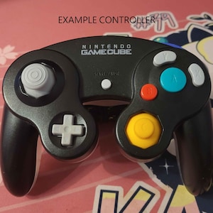 Op de afbeelding: Zwarte Nintendo GameCube-controller met grijze, blauwe, rode en gele knoppen. De controller heeft een grijze joystick en een witte D-pad. De tekst "EXAMPLE CONTROLLER" staat boven de controller.