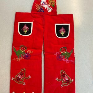 May include: Red embroidered apron with a bird and floral design on the bib. The apron features embroidered green creatures and frogs on the legs, with black and white trim. The apron has a cream-colored handle.