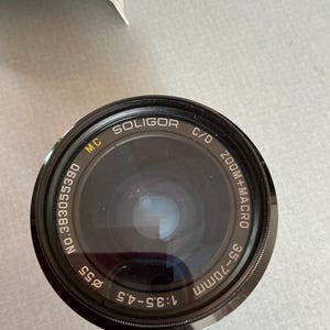 K&ouml;nnte beinhalten: Ein schwarzes Soligor 35-70mm Zoomobjektiv mit Makrofunktion. Das Objektiv hat einen Durchmesser von 55 mm und eine Blende von f/3,5-4,5. Der Text "MC SOLIGOR C/D" ist auf dem Objektiv aufgedruckt.