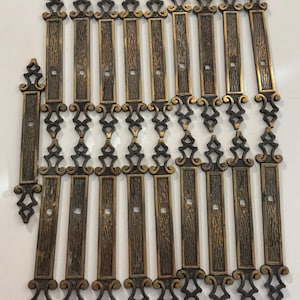 May include: A collection of seventeen elongated metal drawer pulls featuring a dark bronze finish with gold-toned highlights. Each pull has a textured rectangular center panel and ornate, scroll-like decorative ends with a single square mounting hole.