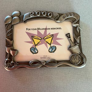 May include: Silver-toned metal photo frame with the years 1999 and 2000. The frame has decorative elements like balloons, champagne glasses, and a clock. The frame is designed to hold a 4x6 inch photo and has the text "For your Millennium memories."