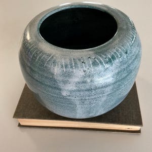 May include: A spherical ceramic vase with a mottled teal and white glaze finish. The exterior features subtle vertical etched lines around the upper shoulder, while the interior is a solid dark black. The vase rests on a rectangular book.