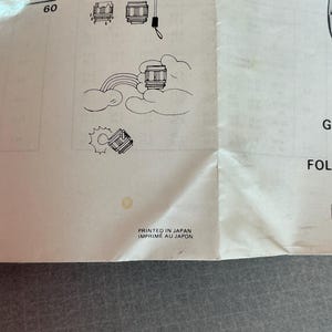 K&ouml;nnte beinhalten: Ein wei&szlig;es Papier mit Illustrationen einer Kameralinse, eines Regenschirms und eines Regenbogens. Der Text "PRINTED IN JAPAN IMPRIME AU JAPON" ist unten auf dem Papier gedruckt.