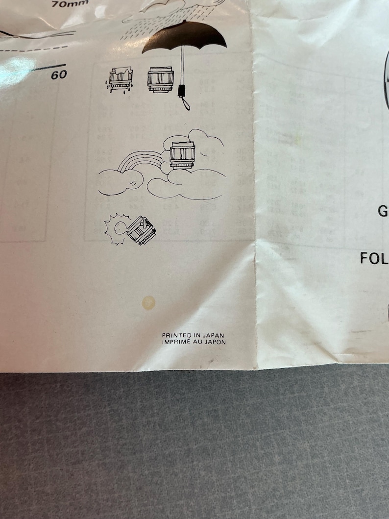 K&ouml;nnte beinhalten: Ein wei&szlig;es Papier mit Illustrationen einer Kameralinse, eines Regenschirms und eines Regenbogens. Der Text "PRINTED IN JAPAN IMPRIME AU JAPON" ist unten auf dem Papier gedruckt.