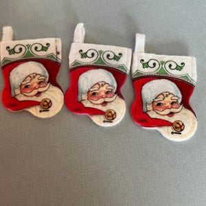 May include: Three small Christmas stockings with a Santa Claus design. Each stocking is red with a white cuff and a green and white decorative border. Santa's face is in the center, with a white beard and a smiling expression.