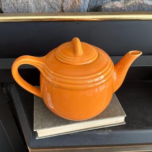 May include: A bright orange ceramic teapot with a matching lid, featuring a rounded body, a curved handle, and a tapered spout. The teapot has a smooth, glossy finish and is designed with concentric circular ridges around the lid and the upper body.
