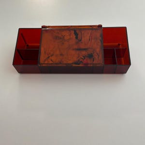 May include: A rectangular, translucent red acrylic organizer box featuring a hinged lid with a marbled brown and black pattern. The interior is divided into multiple compartments, with open sections on the left and right sides and a central lidded area.