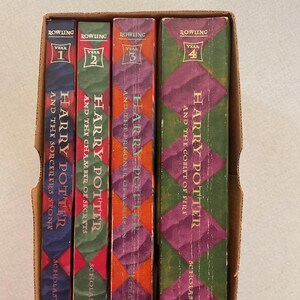 May include: A set of four hardcover books in a cardboard box. The books are all Harry Potter books, with the first four books in the series. The books are all in the same style, with a green and red checkered pattern on the cover. The spines of the books are all red, with the title and author's name in gold lettering. The books are all in good condition.