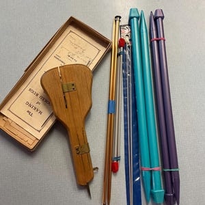 May include: A collection of vintage rug-making tools, including a wooden handle tool, knitting needles in various sizes and colors, and a box with printed instructions. The needles are teal, purple, and gold.