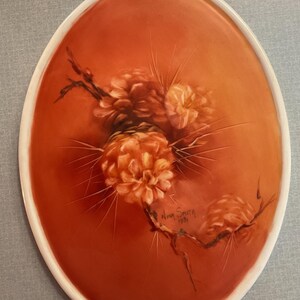 May include: Oval-shaped decorative plate with a white rim and a warm orange center. The center features a floral design in shades of orange and brown, with delicate lines and details. Signed "Nina Smith 1981".