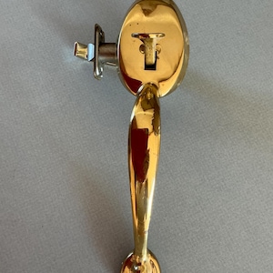 May include: A polished brass door handle with an oval backplate and a long, curved handle. The handle has a keyhole and a latch mechanism. The hardware is a bright gold color and is set against a gray background.