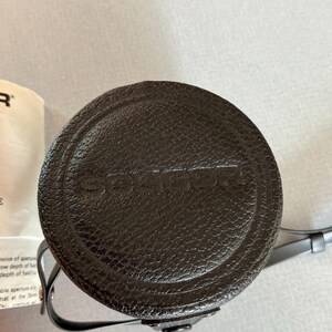 K&ouml;nnte beinhalten: Eine dunkelbraune, kreisf&ouml;rmige Kamerobjektivtasche mit dem eingepr&auml;gten Markennamen "SOLIGOR" auf der Oberseite. Die Tasche hat eine strukturierte Oberfl&auml;che und einen d&uuml;nnen, grauen Riemen. Die Tasche ist wahrscheinlich aus Leder oder einem leder&auml;hnlichen Material.