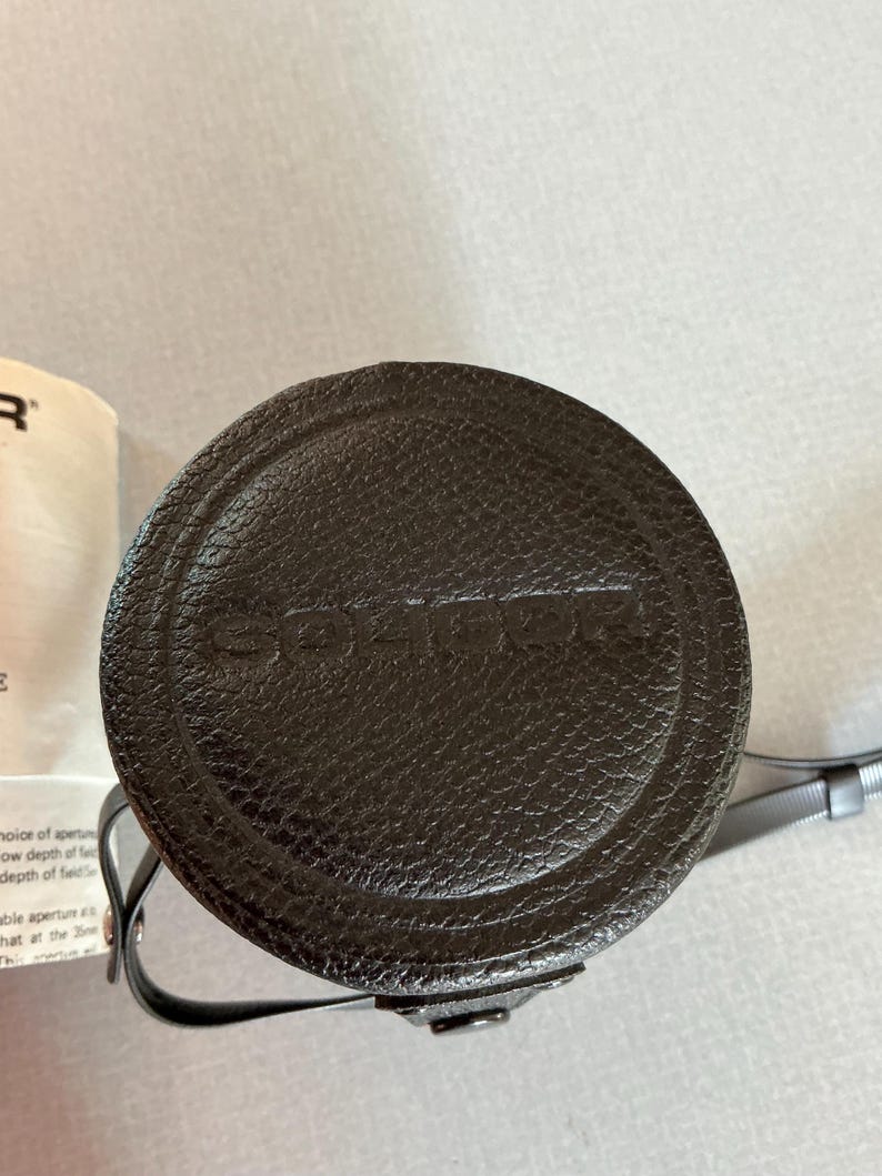 K&ouml;nnte beinhalten: Eine dunkelbraune, kreisf&ouml;rmige Kamerobjektivtasche mit dem eingepr&auml;gten Markennamen "SOLIGOR" auf der Oberseite. Die Tasche hat eine strukturierte Oberfl&auml;che und einen d&uuml;nnen, grauen Riemen. Die Tasche ist wahrscheinlich aus Leder oder einem leder&auml;hnlichen Material.