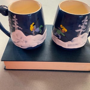 May include: Two blue ceramic mugs with white and pink snow scene designs. Each mug features a skier in green and yellow clothing.