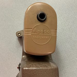 May include: A vintage pencil sharpener with a tan plastic top and a metallic base. The text "Apsco Sharpener" is printed on the top. The sharpener has a crank and a hole for inserting a pencil.