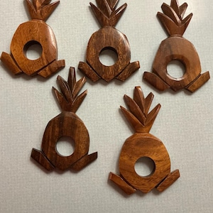 May include: Five wooden pineapple-shaped napkin rings. Each ring is carved from a medium-toned wood, featuring a central circular opening and a detailed pineapple crown. The base of each ring has a small, angular design.