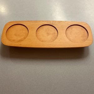 May include: A rectangular wooden tray with three circular indentations. The tray is made of light-colored wood and has a smooth finish. The circular indentations are evenly spaced across the tray's surface. The tray is designed to hold small items.