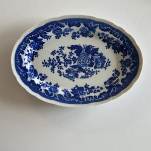 May include: An oval ceramic platter featuring a white base with a deep blue floral pattern. The design includes a central bouquet of flowers and a decorative border of scrolling leaves and blossoms, finished with a ribbed, scalloped edge.