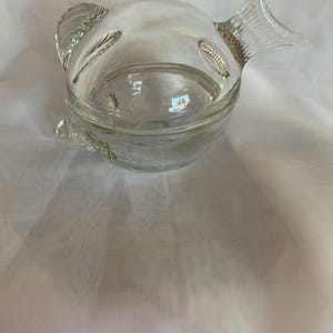 May include: A clear glass fish-shaped container with a detailed tail and fins. The container has a wide opening and a rounded body. The glass is transparent, allowing a view of the contents. The fish-shaped container is likely for decorative or functional use.