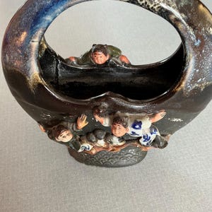 May include: A dark brown ceramic basket with a large handle. The rim has a blue and cream glaze. The basket is decorated with small figurines of people climbing on the side. The figures have painted details.