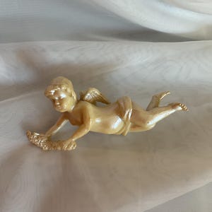 May include: A small, ivory-colored cherub figurine, lying down with outstretched arms and small wings. The cherub holds a garland of flowers. The figurine is set against a soft, white, draped background.