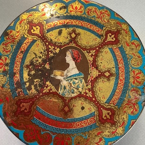 May include: Round vintage tin with a detailed design in gold, red, and blue. The center features a portrait of a woman holding a cake. The tin has an ornate, decorative pattern.